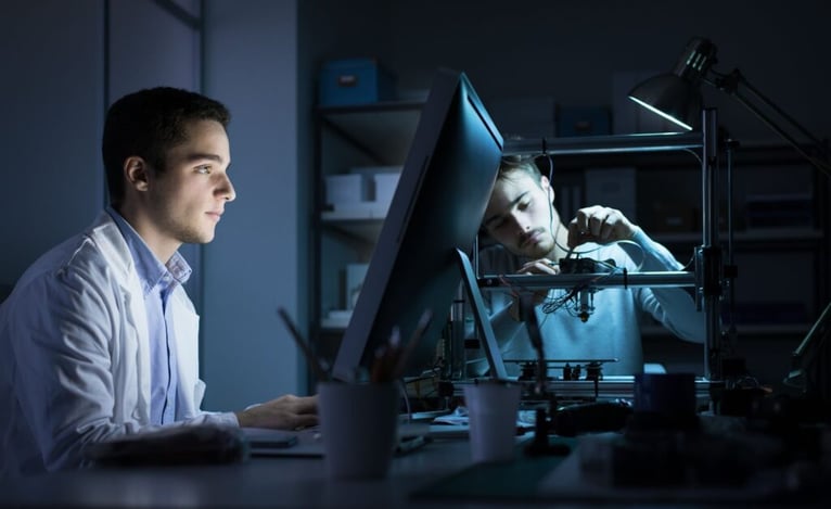 Young male engineer in a lab coat intently works on a computer. Another man, focused under a desk lamp, adjusts a 3D printer.