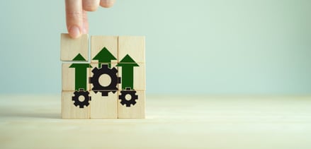 Hand assembling wooden blocks with upward arrows and gear icons, symbolizing growth, productivity, and operational efficiency  