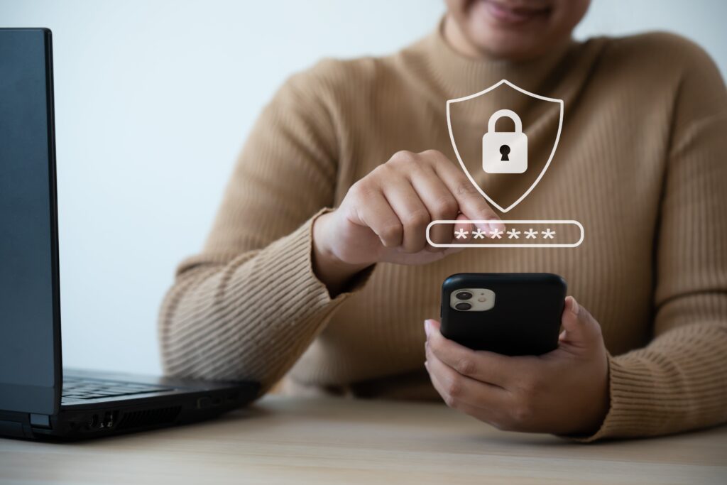 A person in a brown sweater uses a smartphone, with a laptop nearby using MFA to login