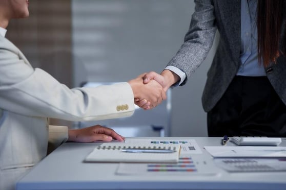 Two business professionals shaking hands across a desk with documents and charts, symbolizing partnership.